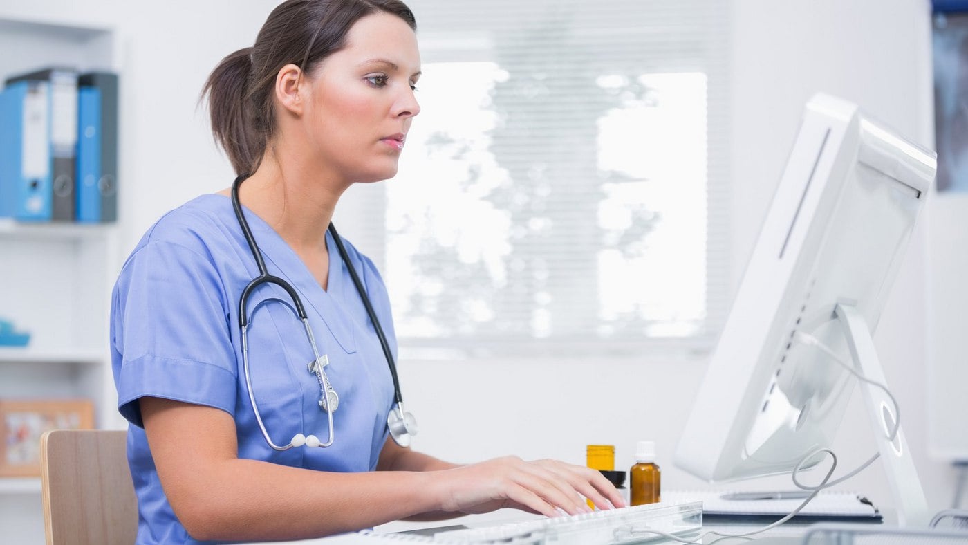 Female healthcare professional in scrubs typing on a keyboard at a computer in a medical office.
