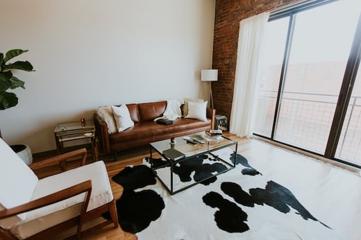 white and brown room with large  sliding door, black and white cow print rug, glass coffee table, and brown leather mid century modern sofa