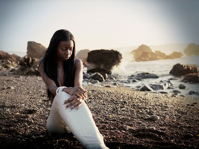 Contemplative young African American woman dressed as a mermaid sitting alone on a rocky beach near the water.