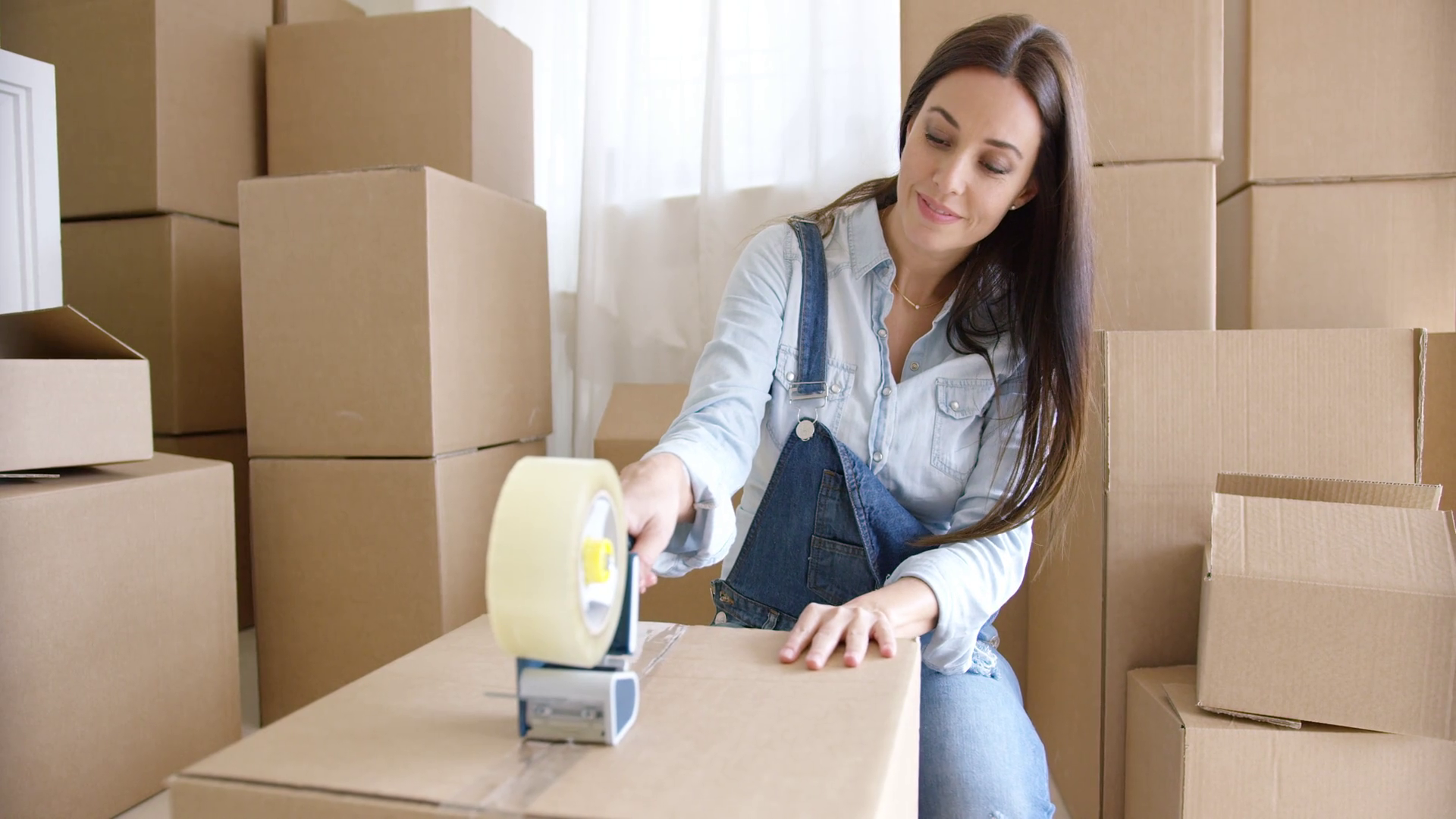 young-woman-moving-home-packing-brown-cardboard-boxes-with-a-roll-of-tape-in-a-close-up-view-with-stacked-boxes-behind-her_rszantomue_thumbnail-full01