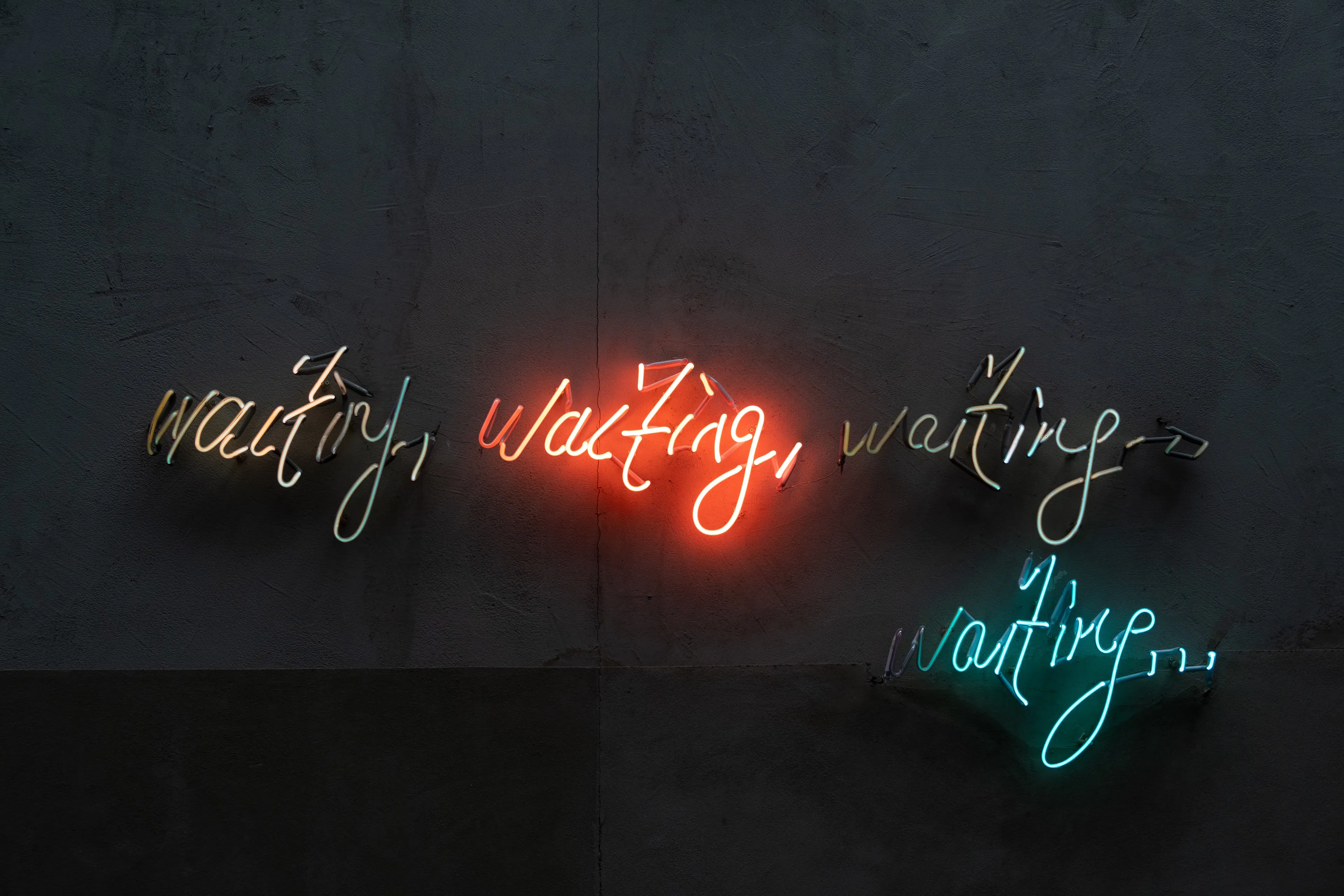 Neon sign on gray wall that says “waiting, waiting, waiting… waiting…”