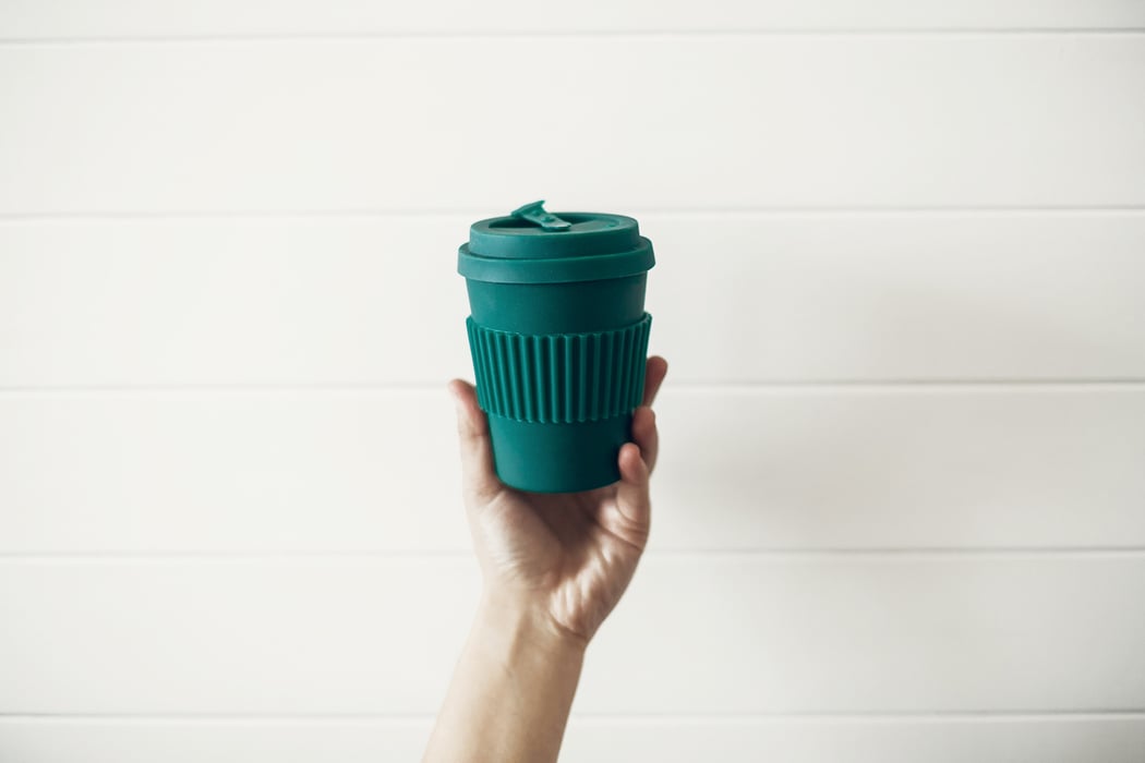 A hand holds up a green reusable coffee mug. 