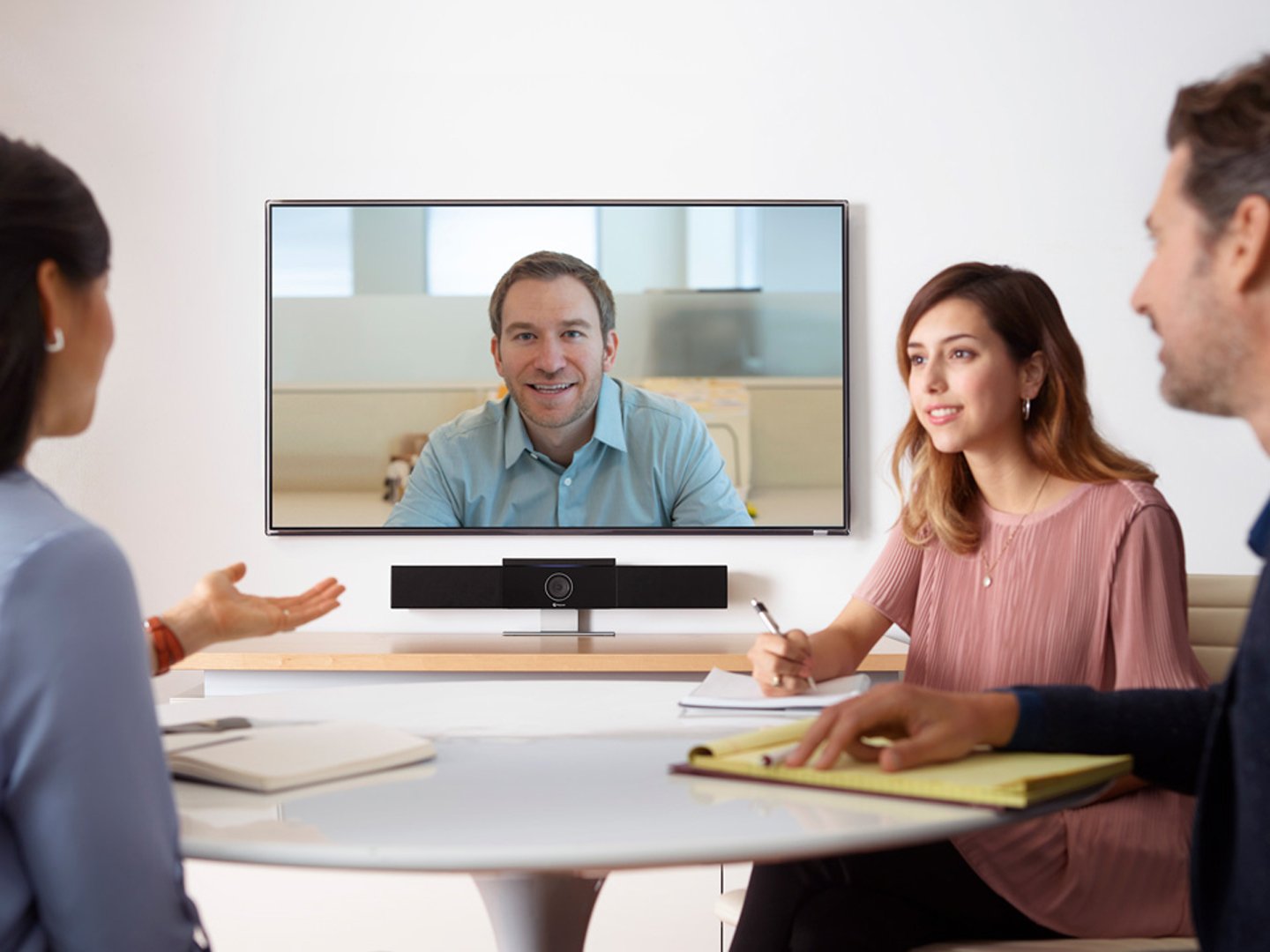 Drei Personen an einem Tisch in einer Sitzung mit einem Display auf dem eine Videokonferenz läuft