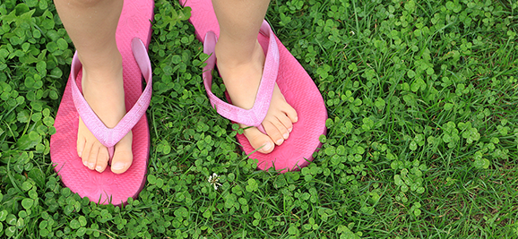 Photograph of child’s feet standing in large adult flip flops over grass and clovers, as an analogy for empathy gained by standing in someone else’s shoes. 