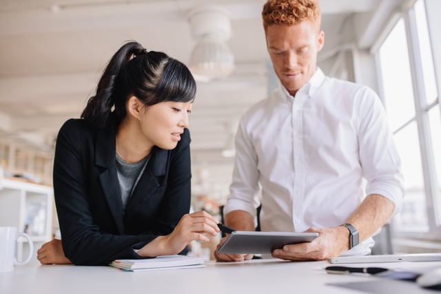 A young man and a young woman standing at an office table, looking at a digital tablet and talking.