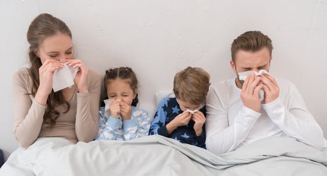 Sick parents and children lying in a bed and blowing noses in napkins.