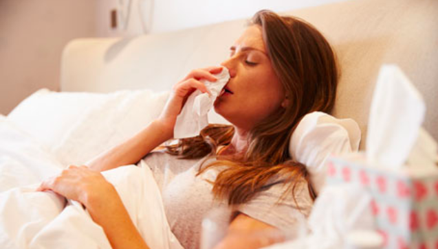 A sick woman blows her nose on a tissue while propped up in bed.
