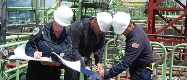 two men and one woman in blue coveralls and white hard hats make notes in a book while checking a piece of equipment.