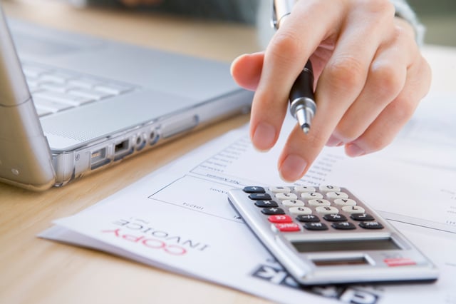 Hand with pen between the fingers typing on a calculator resting on an invoice next to a laptop