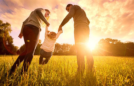 A mother and father lift up a child while walking through a field near sunset