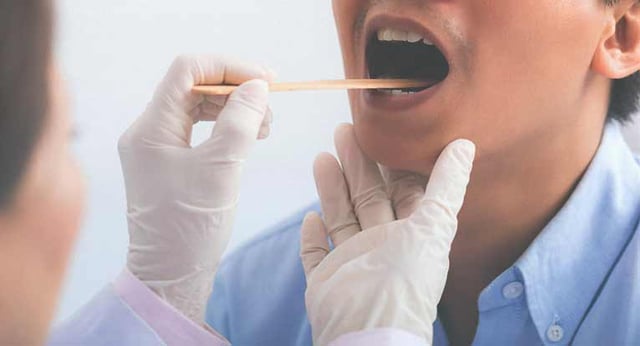 A doctor in a white coat uses a tongue depressor to look at the throat of a man in a blue shirt