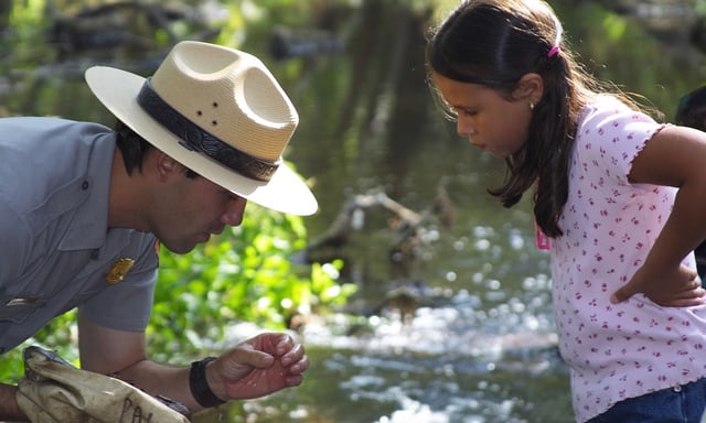 A park ranger shows a young girl a specimen of a plant near a creek.