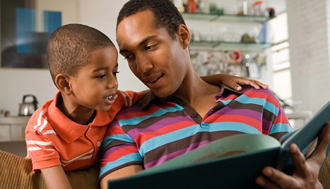 A father and son read a book together. Takes you to  Office of Minority Health's page on men's health awareness month.