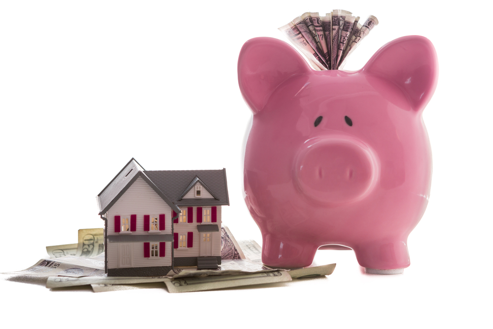 Close up of a pink piggy bank with dollars beside miniature house model on white background