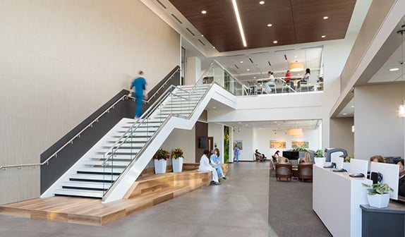 Beachwood Lobby, man in blue scrubs ascending stairs connected to left wall, 2 women talking on large wooden bench below stairs. Across lobby is the check in desk with 2 people manning computers.