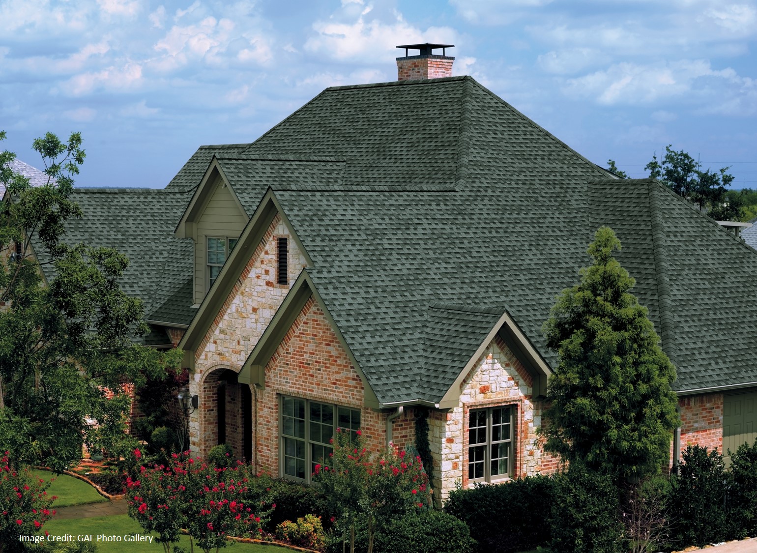 Roof Warranty by GAF A brick home with a green dimensional shingle roof and multiple gables sits behind a landscaped flower garden.