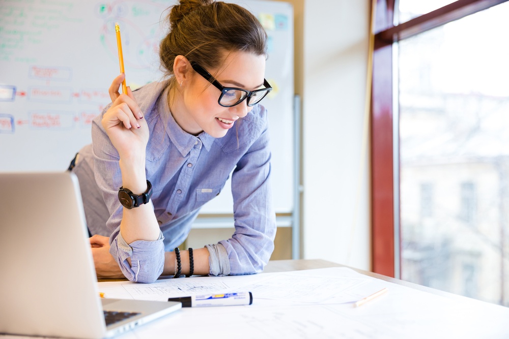 Happy young woman in glasses standing near the window in office and working with blueprint .jpeg