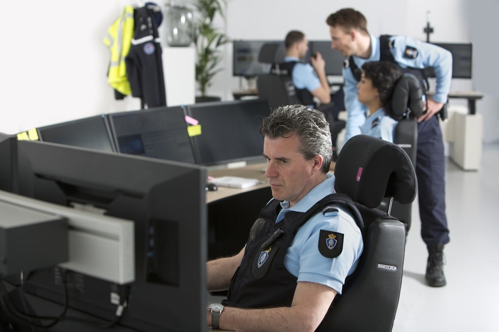 Security guard sitting on Black leather Secur 24 desk chair looking at surveilance monitors with other security guards in the background