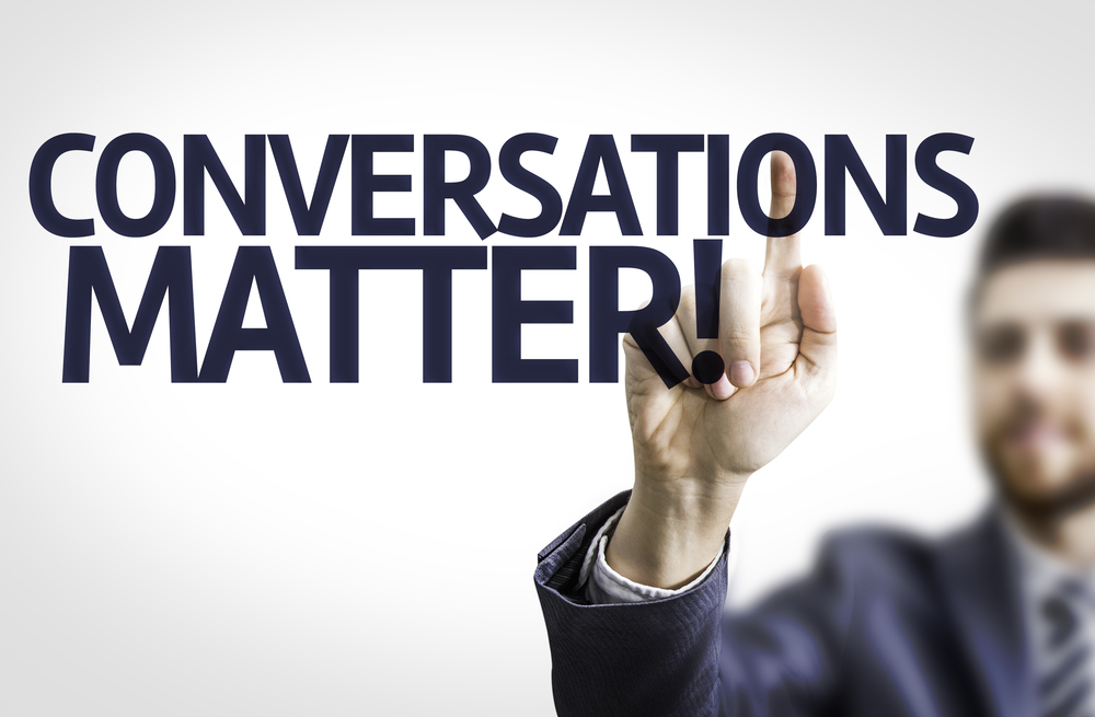 A blurry person in a suit raises their in focus hand to be called on behind ALL CAPS text reading "Conversations Matter!"