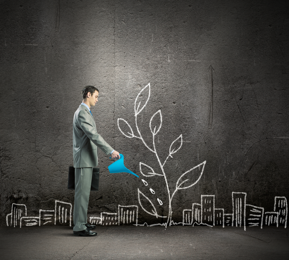 Person in a suit holding a briefcase in one hand and a watering can in the other. The person is "watering" a plant that is part of a chalk drawing cityscape.