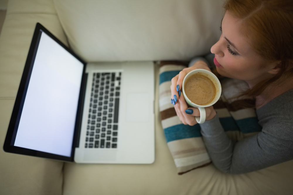 A person is lying on a couch reading a laptop screen while sipping coffee.