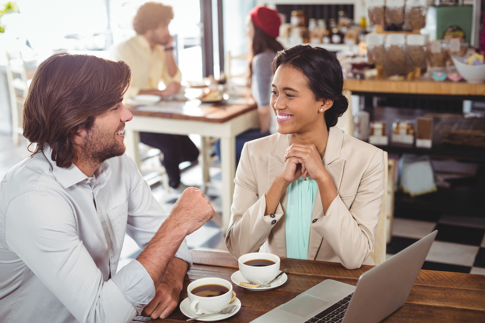 Two colleagues smile while having coffee and a conversation at a cafe. They are looking at each other. This is a common scene of employee engagement.