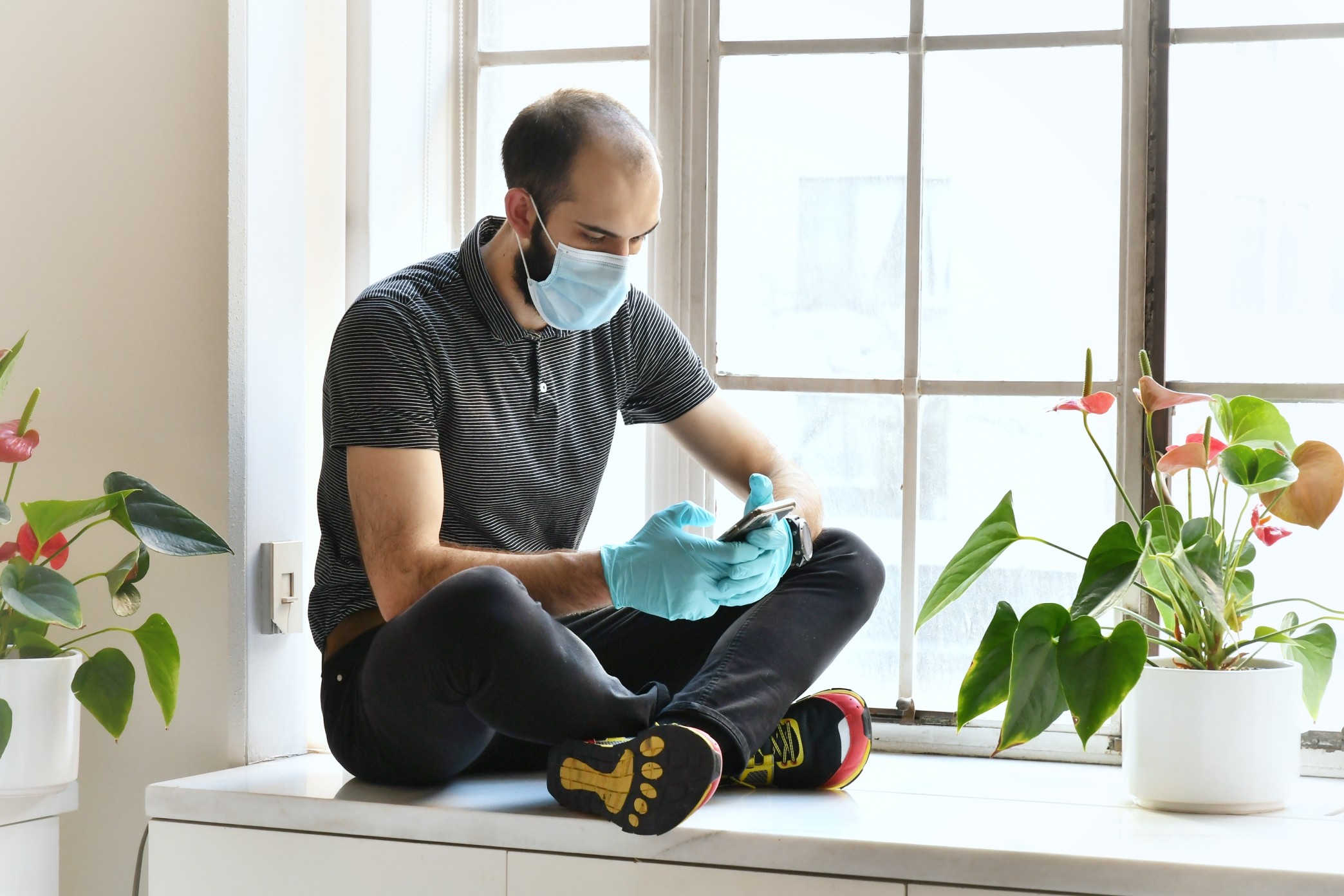 white man sitting on a countertop wearing a face mask and gloves while using a smartphone