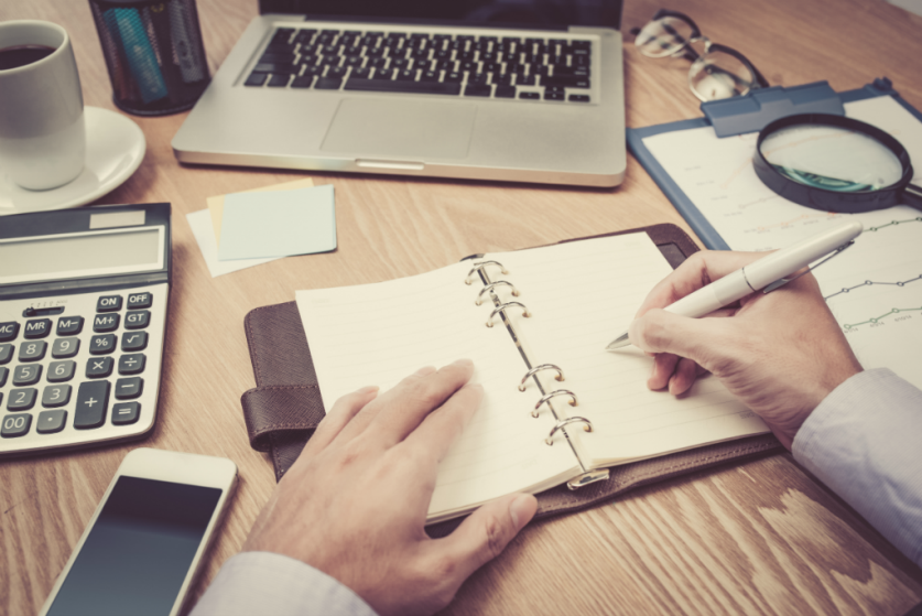 Desk with laptop, calculator, and man writing in planner | SupportingStrategiesFranchise.com