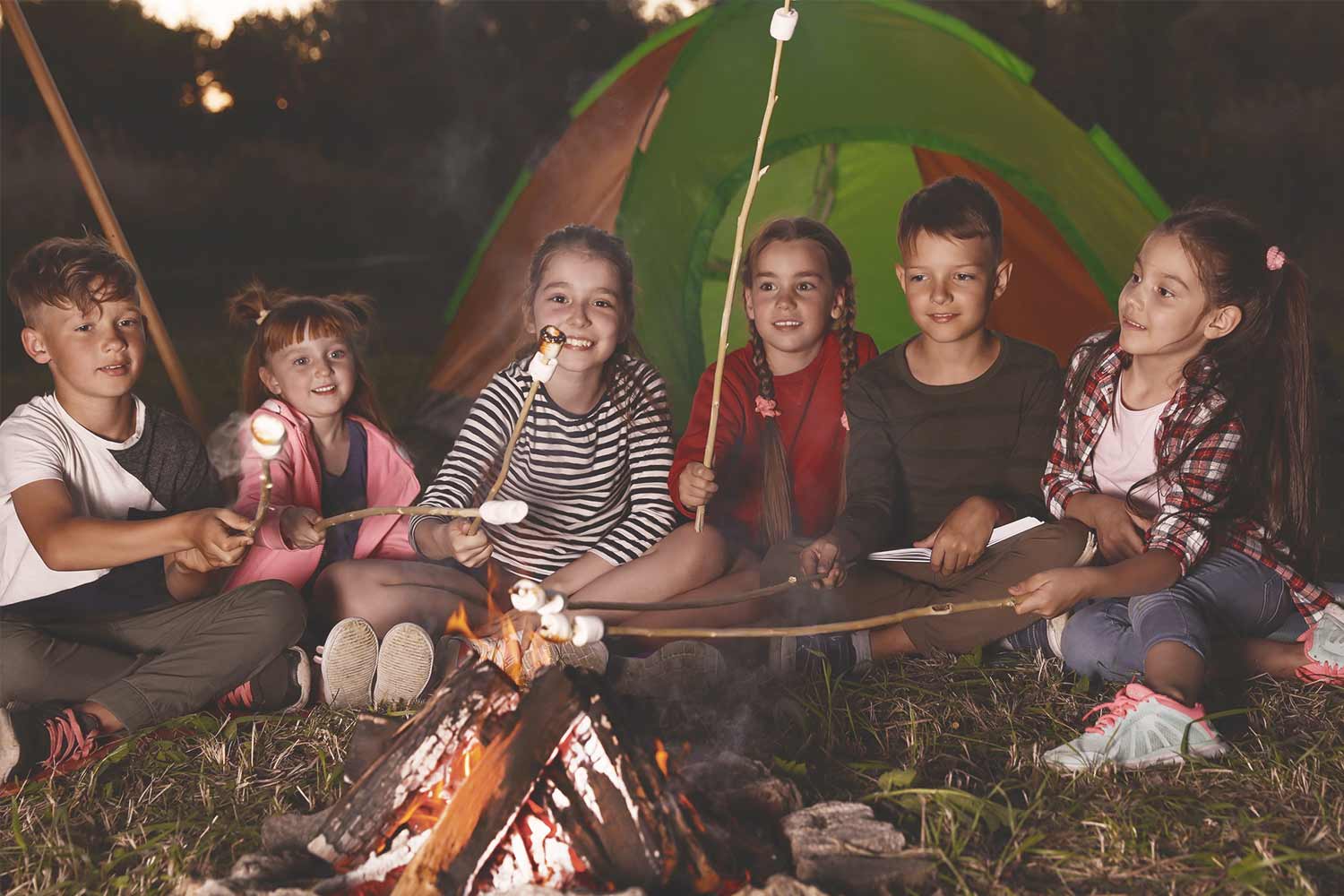 Group of kids camping and roasting marshmallows around a bonfire at night as a summer activity