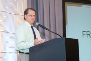 Doug Dwyer stands behind a podium beside a large projector screen as he speaks at the annual DreamMaker Reunion.