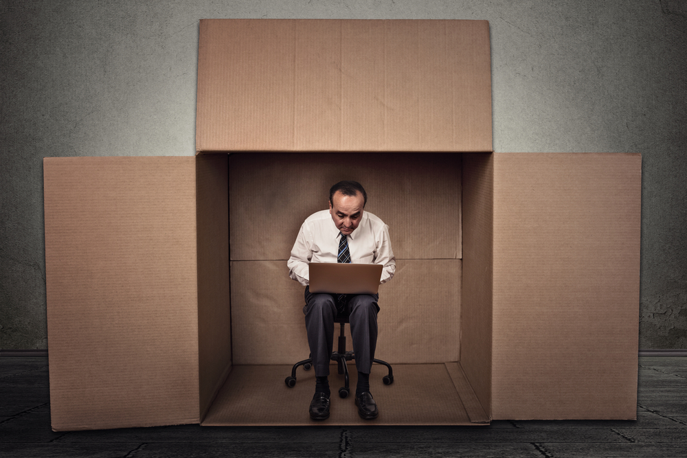 Limitations poor communication of corporate life. Portrait corporate middle aged man working on laptop sitting on chair inside carton box in empty office room