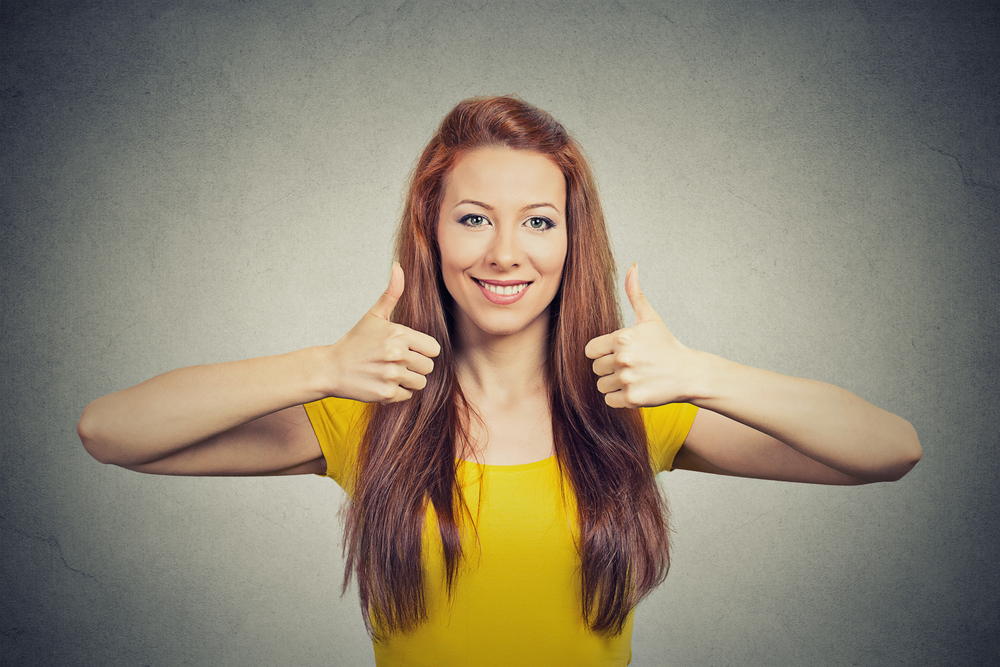 Happy smiling woman with thumbs up gesture isolated grey wall background. Positive emotion face expression sign symbol body language