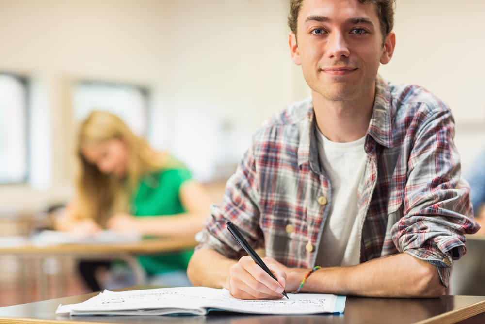 Portrait of a smiling male student with others writing notes in the classroom